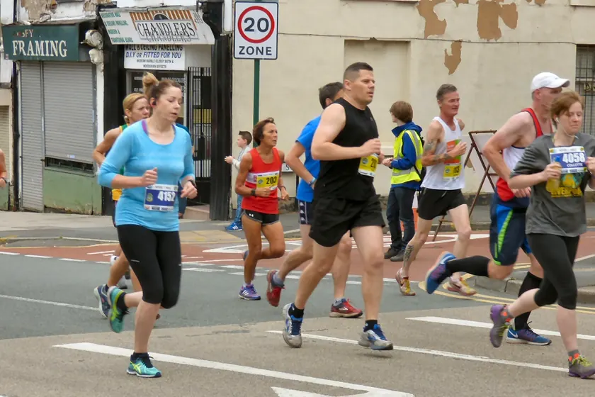 Runners in numbered bibs jogging along a city street past brick storefronts