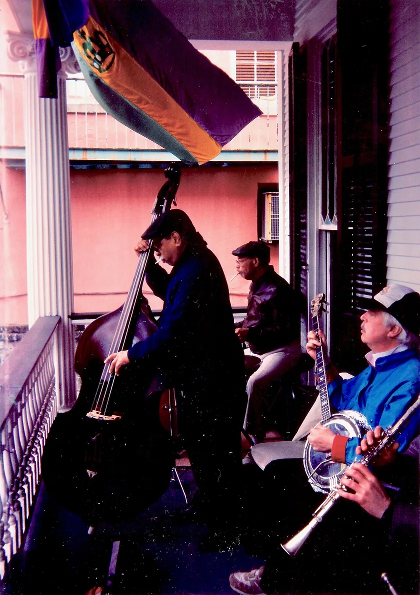 Musicians with brass instruments playing on a porch while a crowd watches from the street