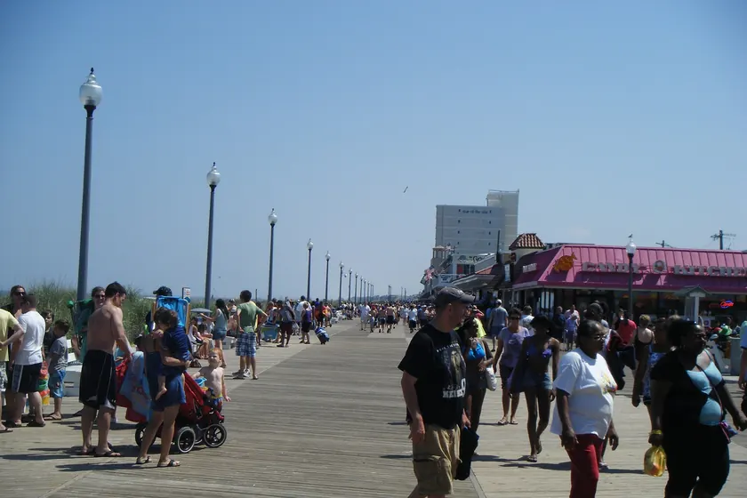 A wooden beach boardwalk lined with shops, with sand on the left and an American flag overhead